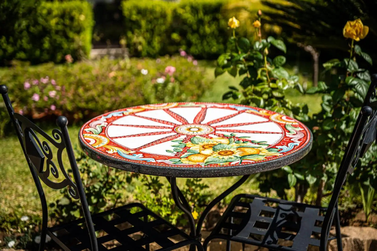 Un tavolo circolare da esterno con un top in piastrelle di ceramica colorate, ornate e dipinte a mano, con un motivo radiale e un motivo floreale, abbinato a due sedie in ferro battuto.
